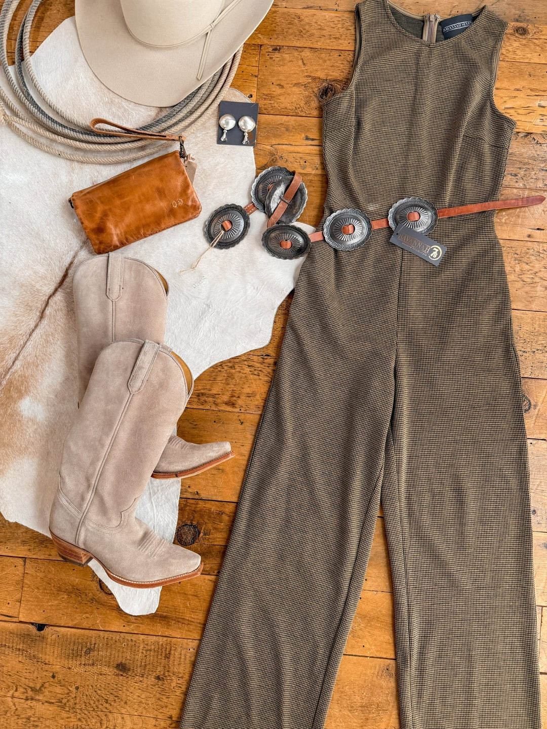 Western-inspired flat lay featuring the Stratford Houndstooth Jumpsuit by Savannah Sevens western life{&}style, beige cowboy boots, brown leather concho belt, matching jewelry, brown clutch, cowboy hat, and coiled rope on a wood floor.