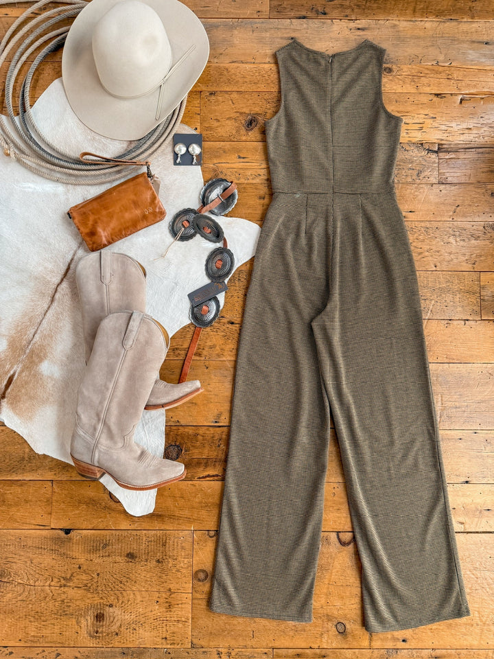 Flat lay of the Stratford Houndstooth Jumpsuit by Savannah Sevens western life{&}style, tan cowboy boots, a wide-brimmed hat, brown clutch, concho belt, and earrings on a wooden floor with a cowhide rug.