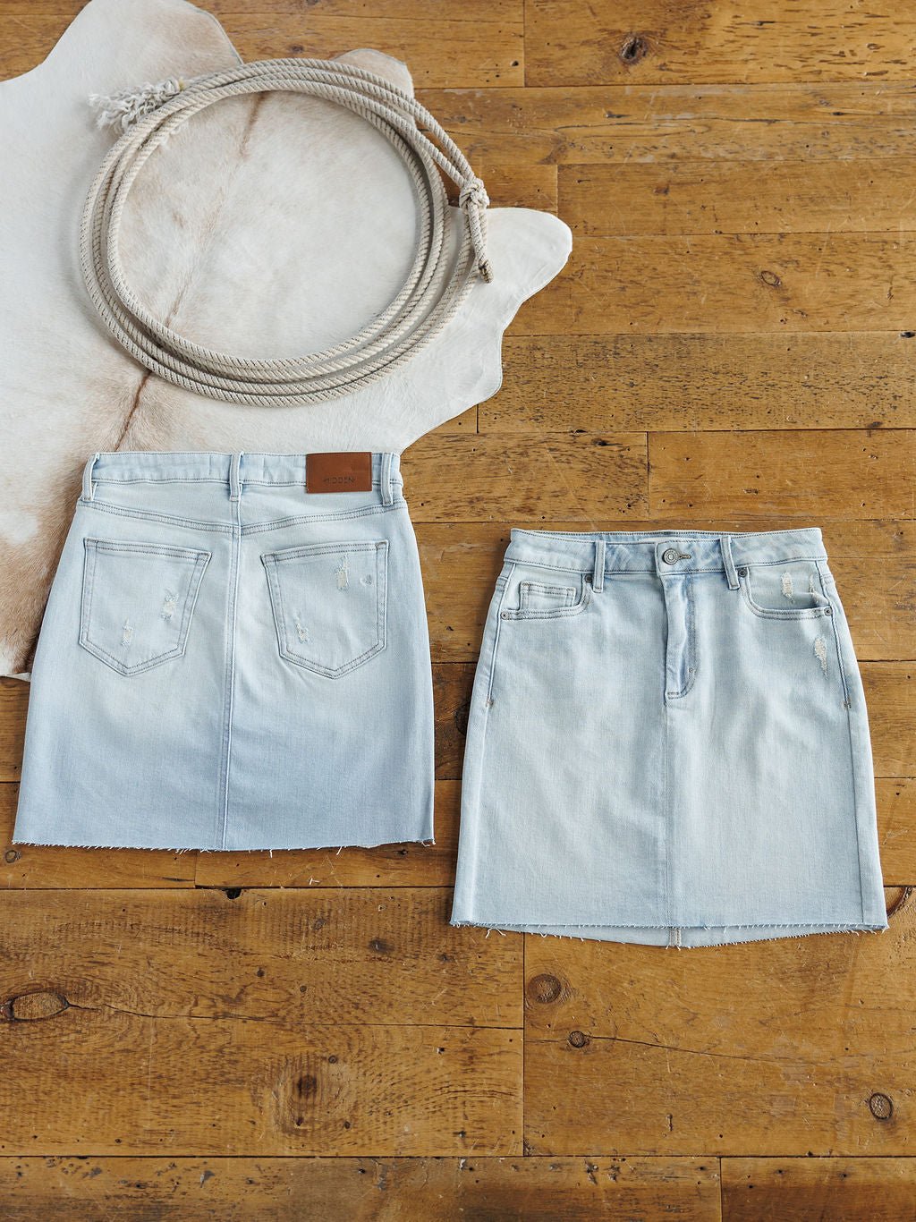 Two The Sumner Skirts by Savannah Sevens western life{&}style are laid flat: one shows the back with pockets and a patch, the other the front with buttons, pockets, and a raw hem. A coiled rope sits on a white animal hide in the corner.