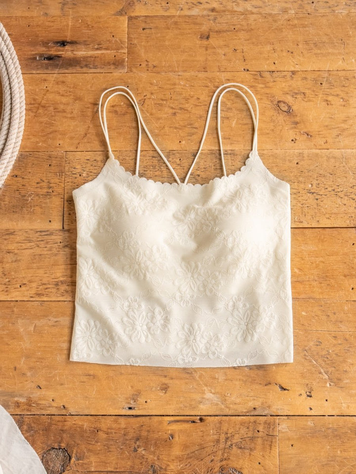 The Surrey Bralette by Savannah Sevens western life{&}style, shown laid flat on a wooden surface, features cream-colored lace with a subtle floral pattern and thin spaghetti straps.