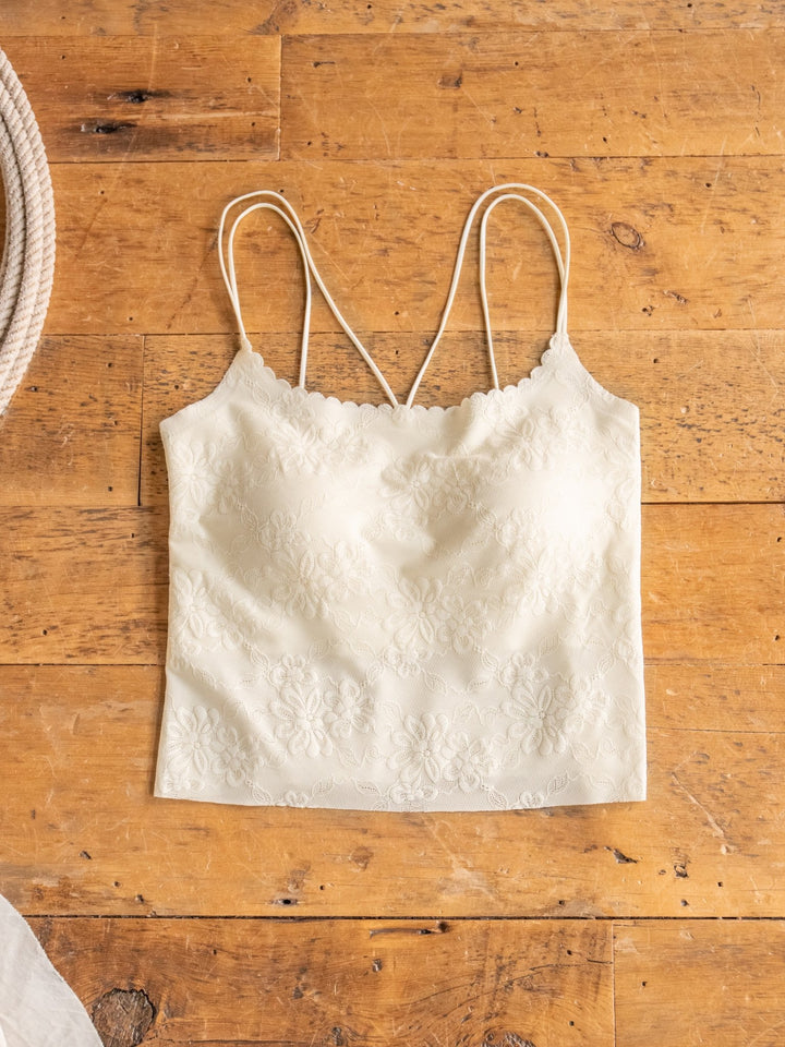 The Surrey Bralette by Savannah Sevens western life{&}style, shown laid flat on a wooden surface, features cream-colored lace with a subtle floral pattern and thin spaghetti straps.