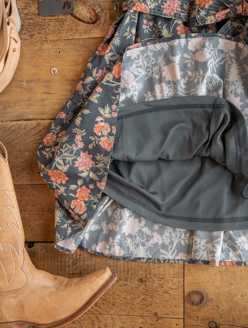 The Sutherland Floral Puff Sleeve Dress by Savannah Sevens western life{&}style, featuring a gray lining, is spread on a wooden floor next to tan cowboy boots and a delicate necklace with a small pendant.