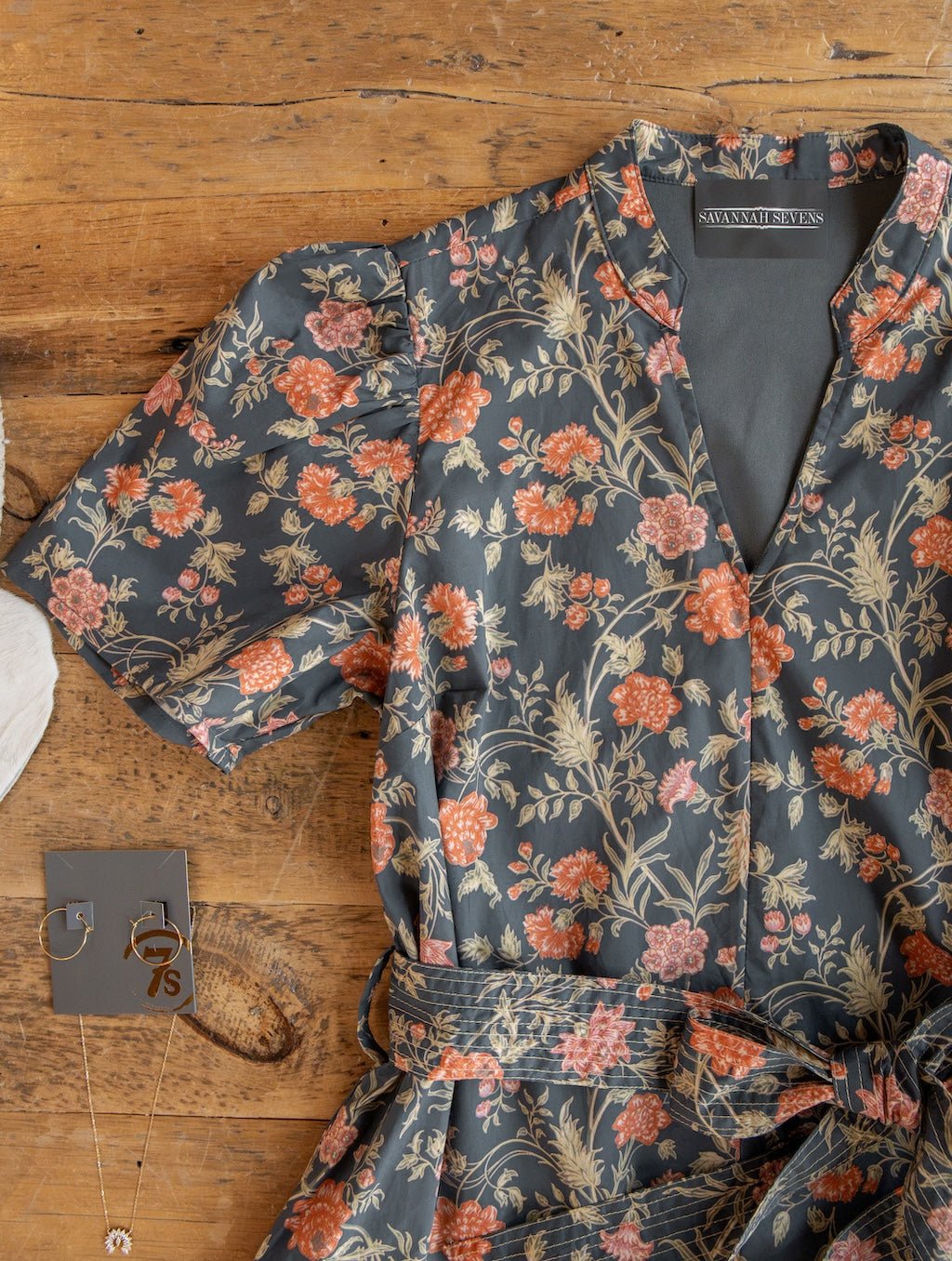 The Sutherland Floral Puff Sleeve Dress by Savannah Sevens western life{&}style, featuring red and beige flowers on a dark background, is displayed on a wooden surface alongside gold jewelry and a necklace.