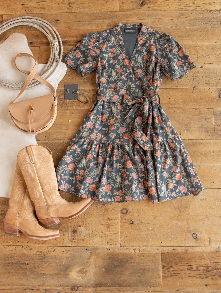 The Sutherland Floral Puff Sleeve Dress by Savannah Sevens western life{&}style is styled with tan cowboy boots, a tan crossbody bag, coiled rope, and earrings, all laid out on a wooden floor.
