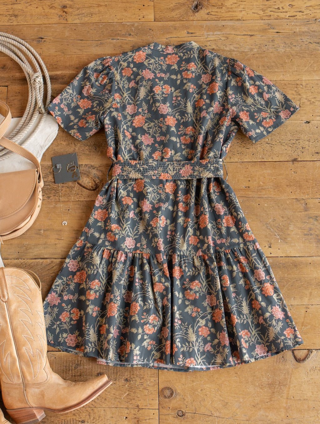 The Sutherland Floral Puff Sleeve Dress by Savannah Sevens western life{&}style, featuring orange and pink flowers on soft cotton, is laid flat on a wooden floor. It’s surrounded by cowboy boots, a tan saddlebag, and silver jewelry.