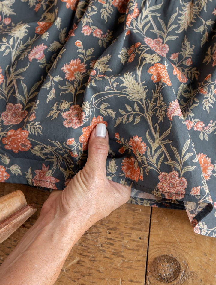 A hand with light nail polish holds the Sutherland Floral Puff Sleeve Dress by Savannah Sevens western life{&}style, featuring a dark fabric with an orange and cream floral pattern, over a wooden surface.