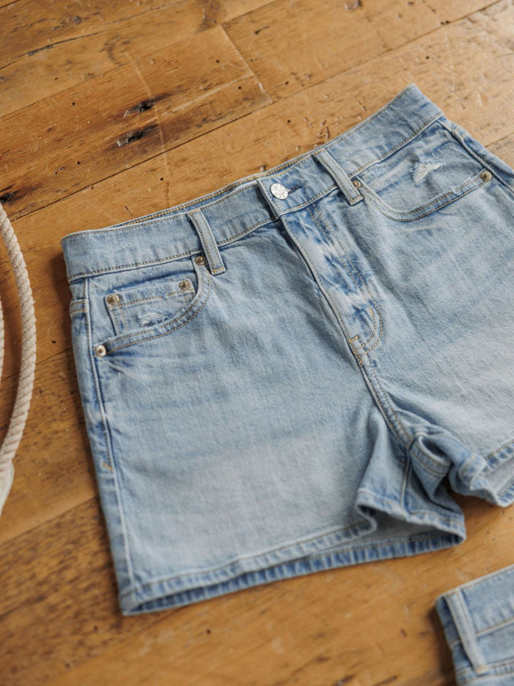 The Sutton Shorts by Savannah Sevens western life{&}style are light blue high rise denim shorts with a medium light wash, front pockets, and button fly, displayed flat on a wooden floor with a rope partially visible on the left.