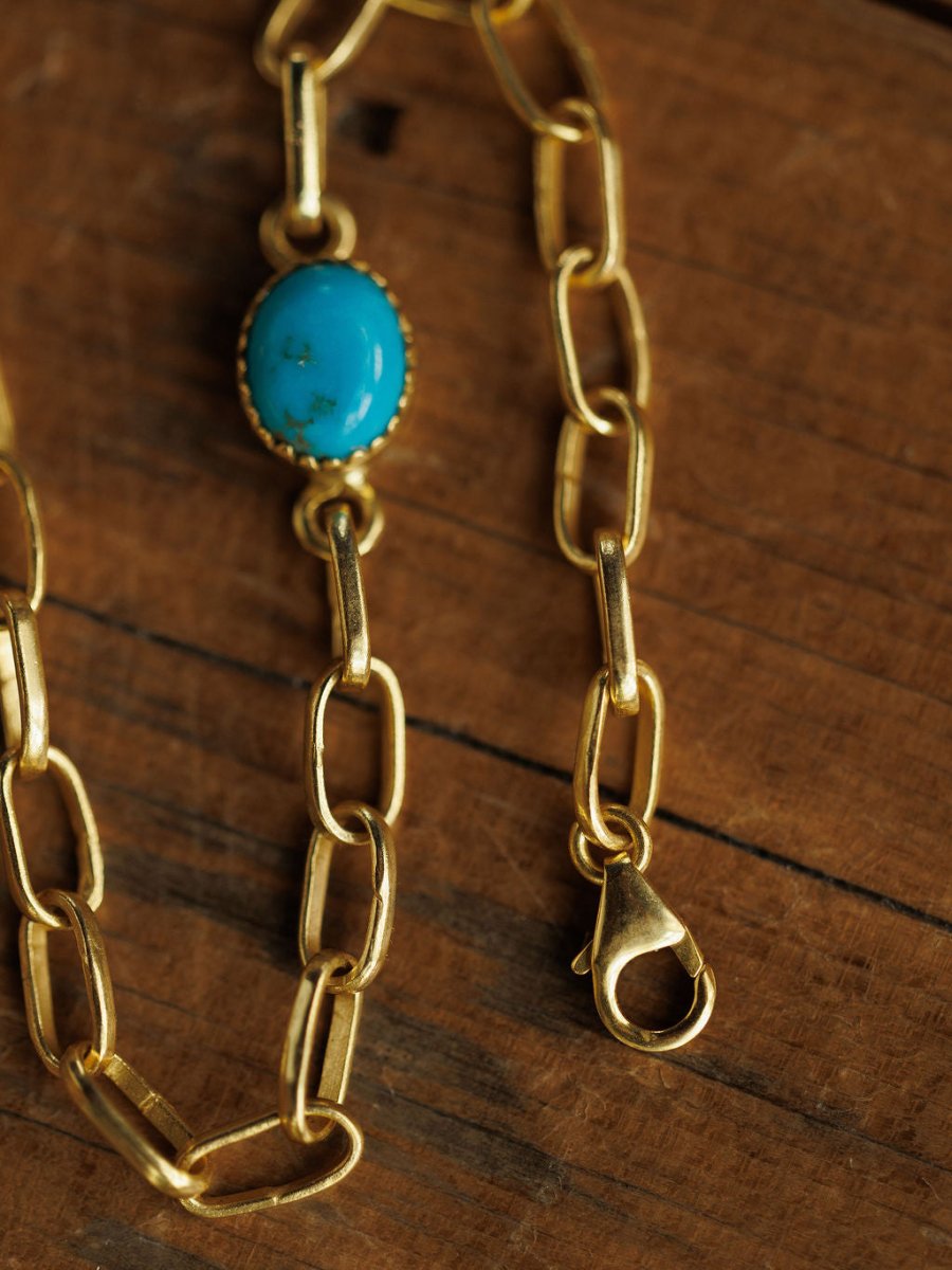 The Suwanee Bracelet by Savannah Sevens western life{&}style features gold-plated sterling silver elongated links and a turquoise oval stone with a lobster clasp, shown on a wooden surface.