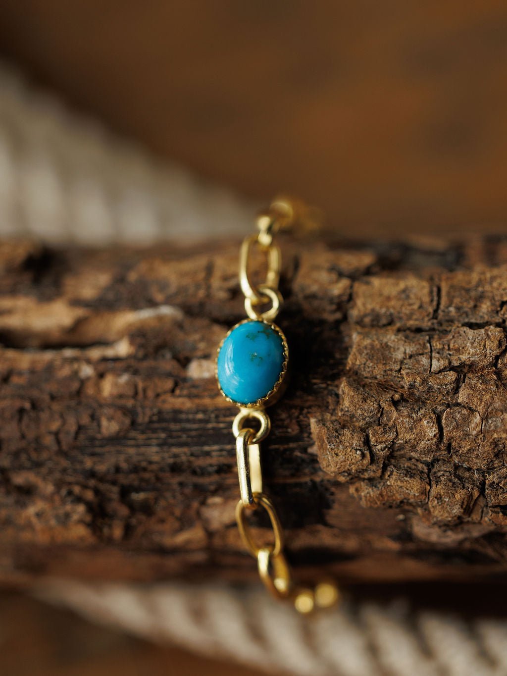 The Suwanee Bracelet by Savannah Sevens western life{&}style features an oval turquoise stone on a gold plated sterling silver chain, displayed on textured brown wood with a blurred coiled rope in the background.