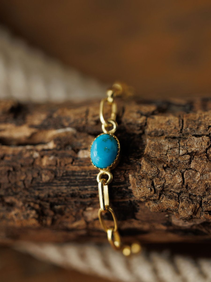 The Suwanee Bracelet by Savannah Sevens western life{&}style features an oval turquoise stone on a gold plated sterling silver chain, displayed on textured brown wood with a blurred coiled rope in the background.