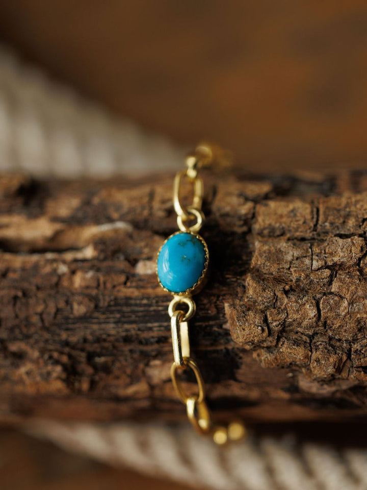 The Suwanee Bracelet by Savannah Sevens western life{&}style features an oval turquoise stone on a gold plated sterling silver chain, displayed on textured brown wood with a blurred coiled rope in the background.