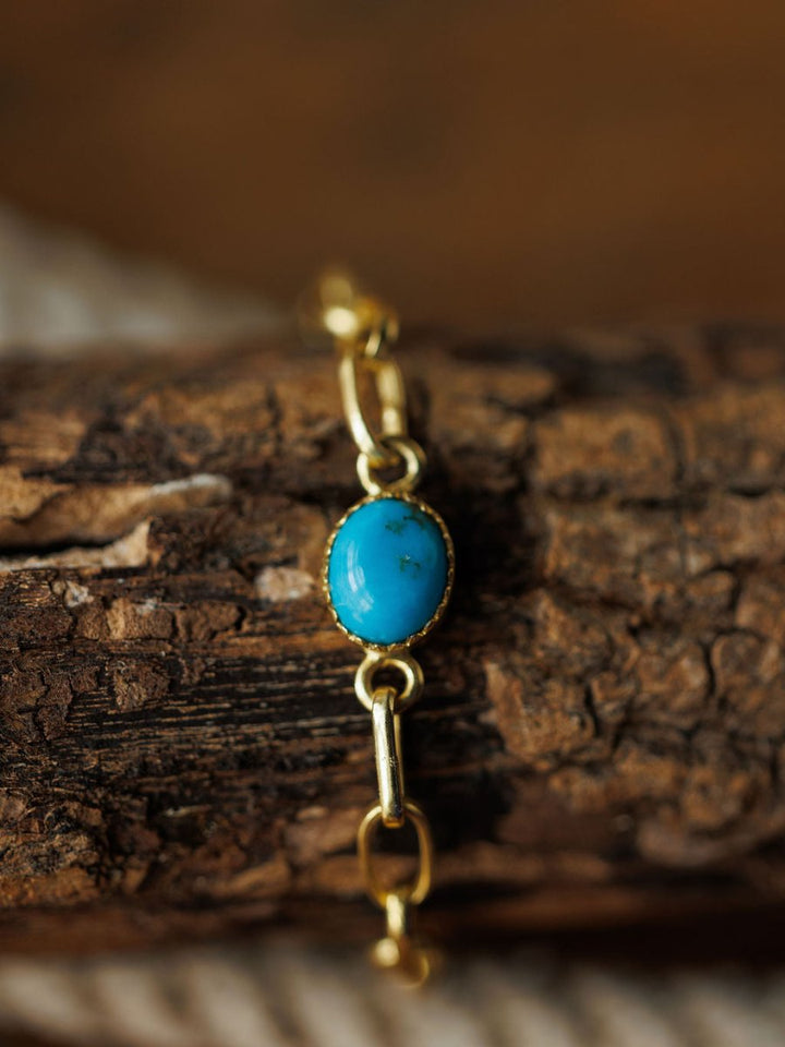 The Suwanee Bracelet by Savannah Sevens western life{&}style is a handmade turquoise oval gemstone paperclip design with gold plated sterling silver links, shown on a rustic wood surface. A striking blue stone accents the center.