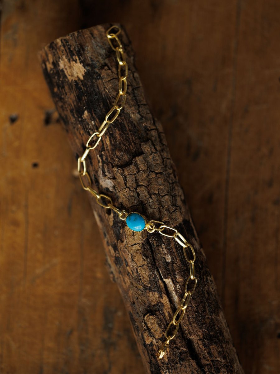 The Suwanee Bracelet by Savannah Sevens western life{&}style features handmade turquoise paperclips, elongated gold-plated sterling silver links, and a bright blue oval stone, shown draped across textured wood with a rustic brown background.
