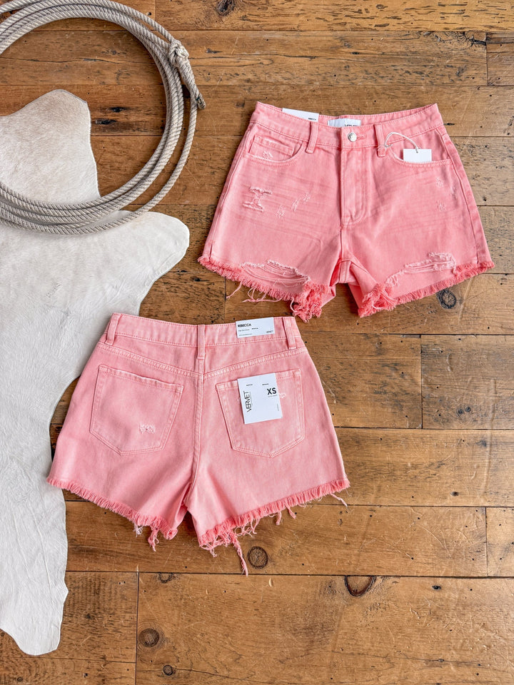 Two pairs of The Swayzi Shorts by Savannah Sevens western life{&}style in high rise salmon pink denim with frayed hems are displayed on a wooden floor, one showing the front, the other the back with visible tags. A coiled rope and white hide rug are nearby.