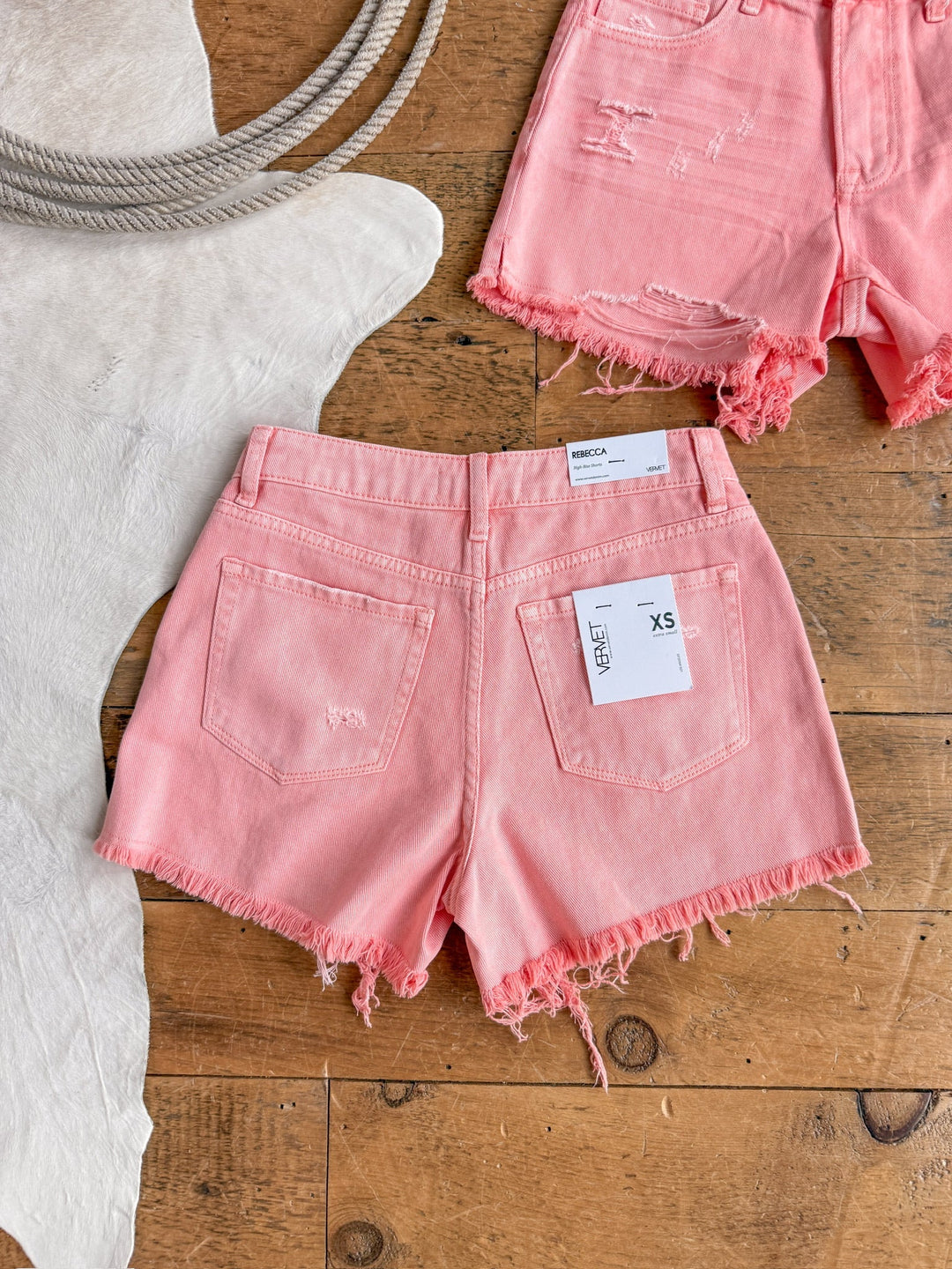 The Swayzi Shorts by Savannah Sevens western life{&}style, in salmon pink denim with frayed hems, are displayed on a wooden floor near a light cowhide rug and rope. A second high rise vintage wash denim short appears in the top right corner.