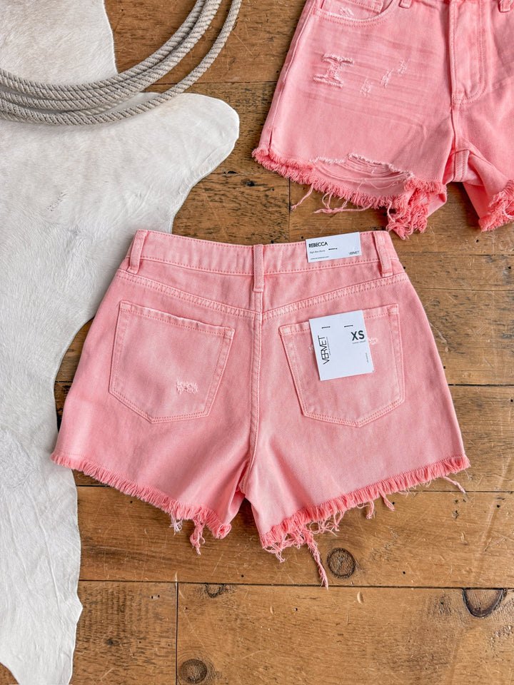 The Swayzi Shorts by Savannah Sevens western life{&}style, in salmon pink denim with frayed hems, are displayed on a wooden floor near a light cowhide rug and rope. A second high rise vintage wash denim short appears in the top right corner.