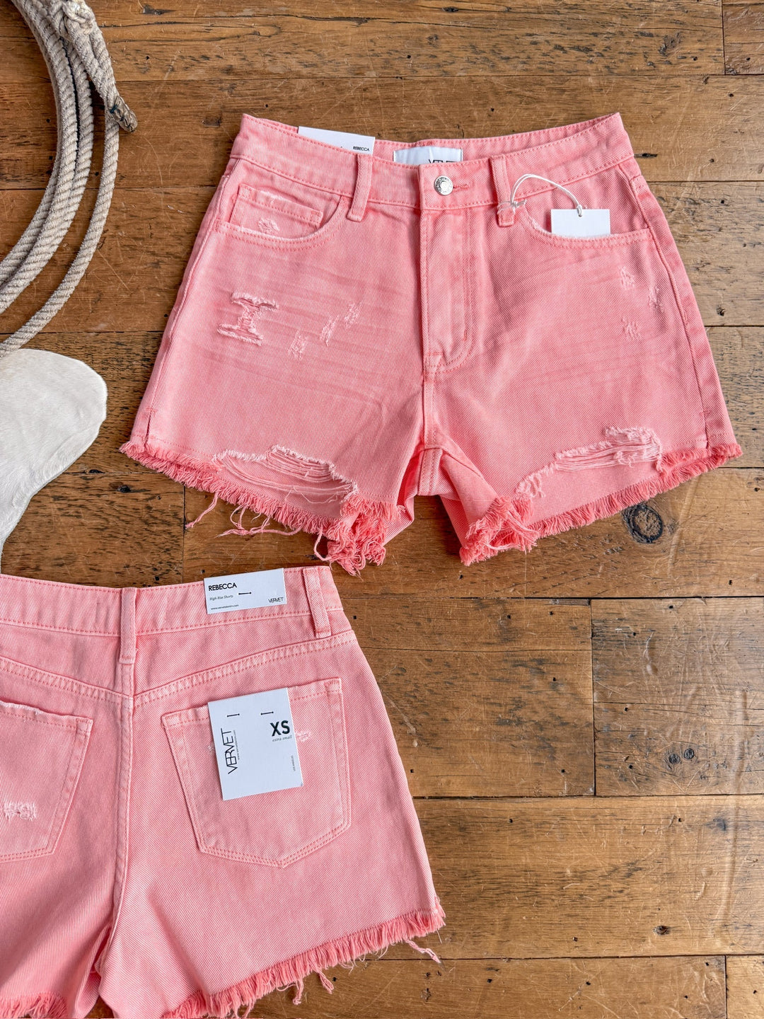 Two pairs of The Swayzi Shorts by Savannah Sevens western life{&}style in salmon pink denim with vintage wash, frayed hems, and distressed details are laid out on a wooden floor—one front view and one back view with tags attached.