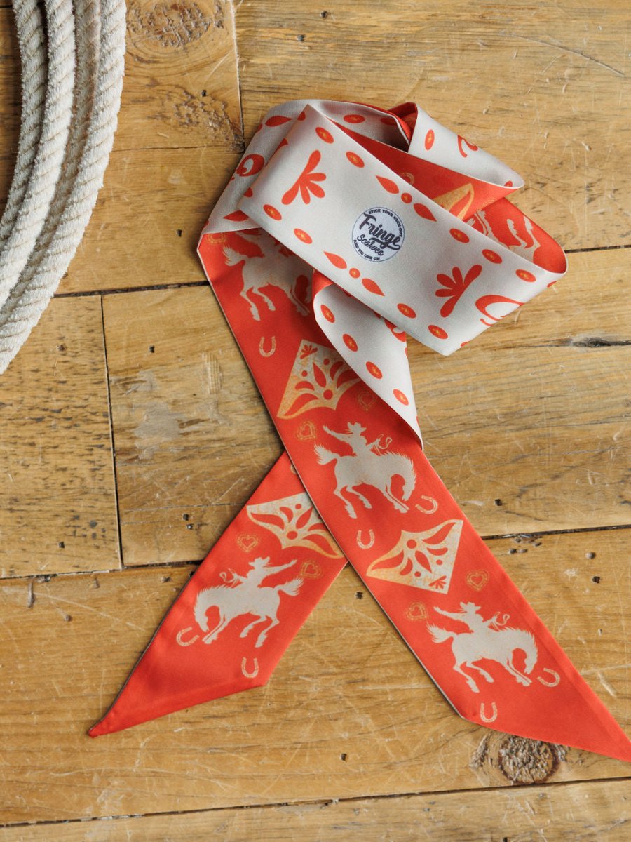 The Sweetheart Twilly by Savannah Sevens western life{&}style, a red and cream poly chiffon cowgirl scarf with horse and horseshoe patterns, rests on wood near a coiled rope. It features western illustrations and a small circular Fringe Scarves logo.