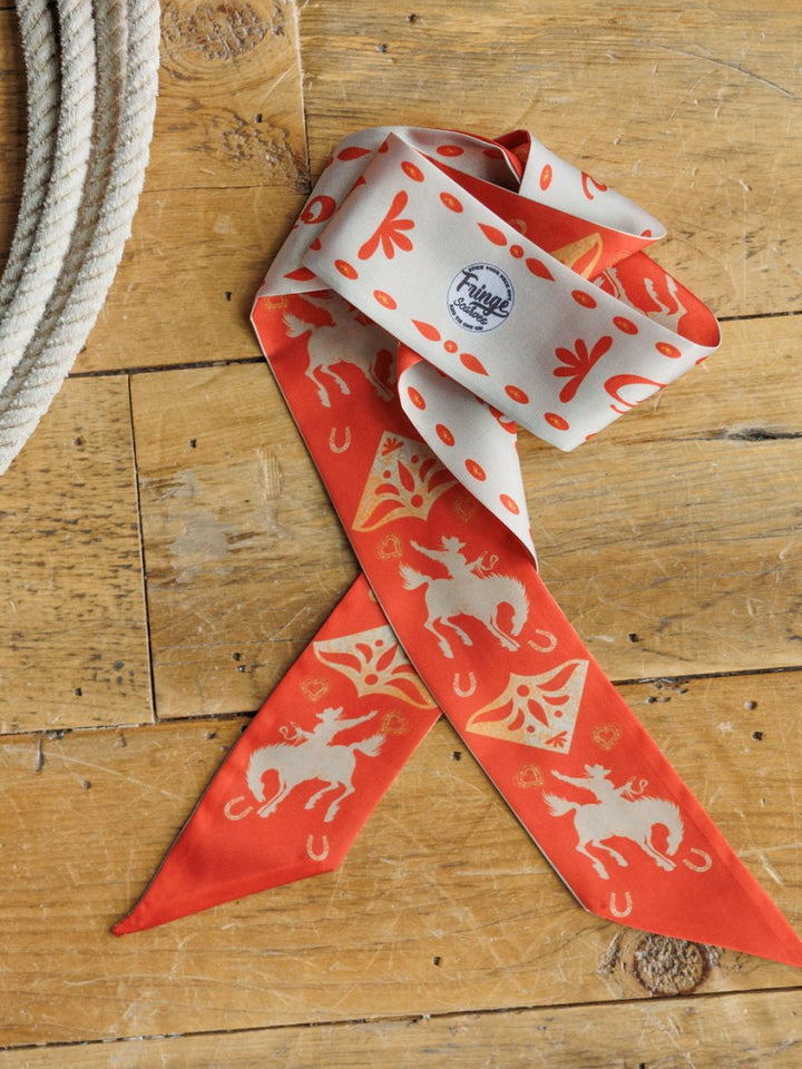 The Sweetheart Twilly by Savannah Sevens western life{&}style, a red and cream poly chiffon cowgirl scarf with horse and horseshoe patterns, rests on wood near a coiled rope. It features western illustrations and a small circular Fringe Scarves logo.
