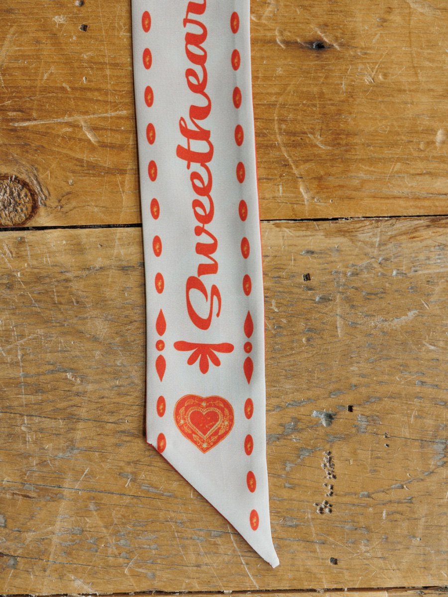 The Sweetheart Twilly by Savannah Sevens western life{&}style features white fabric with red “Sweetheart” text and heart designs, displayed on rustic wood—ideal as a charming accent or styled as a cowgirl scarf.