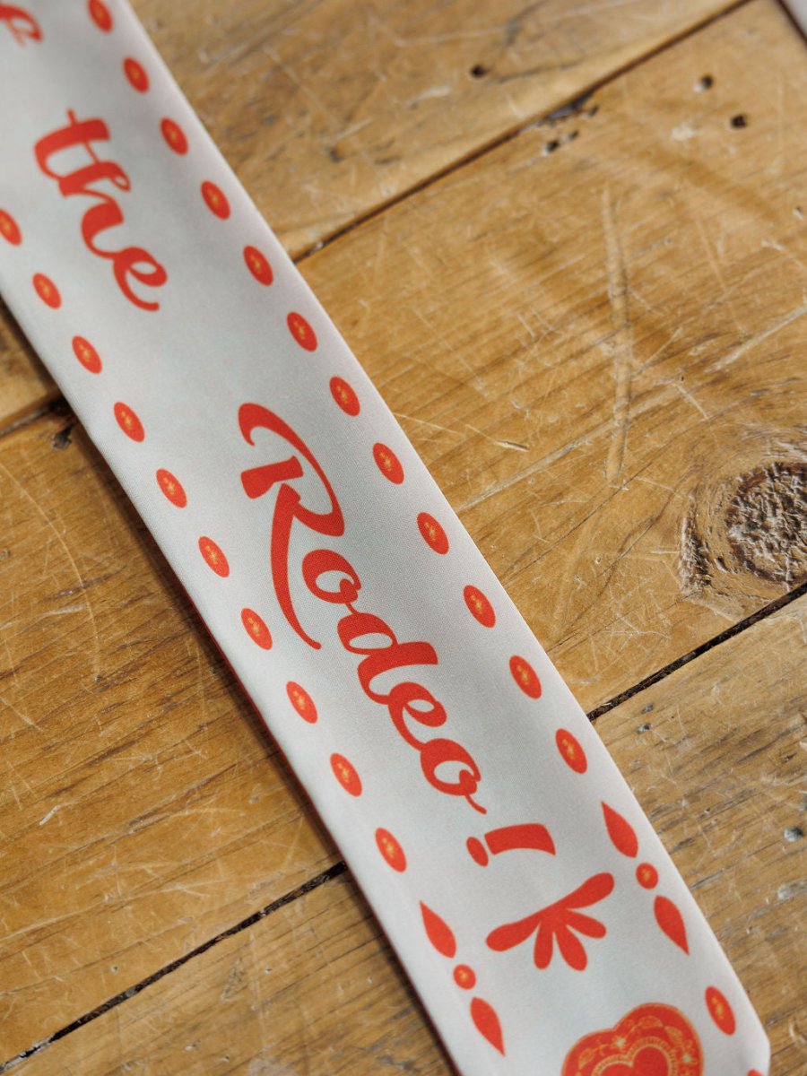 The Savannah Sevens western life{&}style Sweetheart Twilly, a white cowgirl scarf with red dots and “Rodeo!” in bold script, is displayed diagonally on a rustic wooden surface.