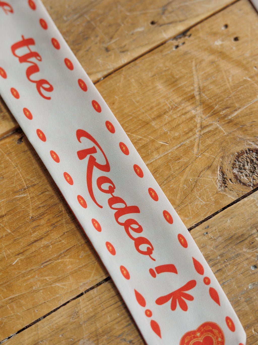The Savannah Sevens western life{&}style Sweetheart Twilly, a white cowgirl scarf with red dots and “Rodeo!” in bold script, is displayed diagonally on a rustic wooden surface.