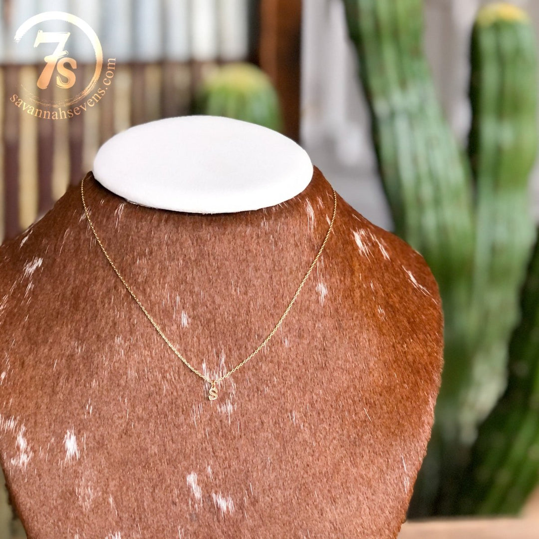 The Sylvan Initial Necklace by Savannah Sevens western life{&}style, featuring a gold "S" pendant, is shown on a cowhide-patterned bust with a blurred green cactus and the Savannah Sevens logo in the background.