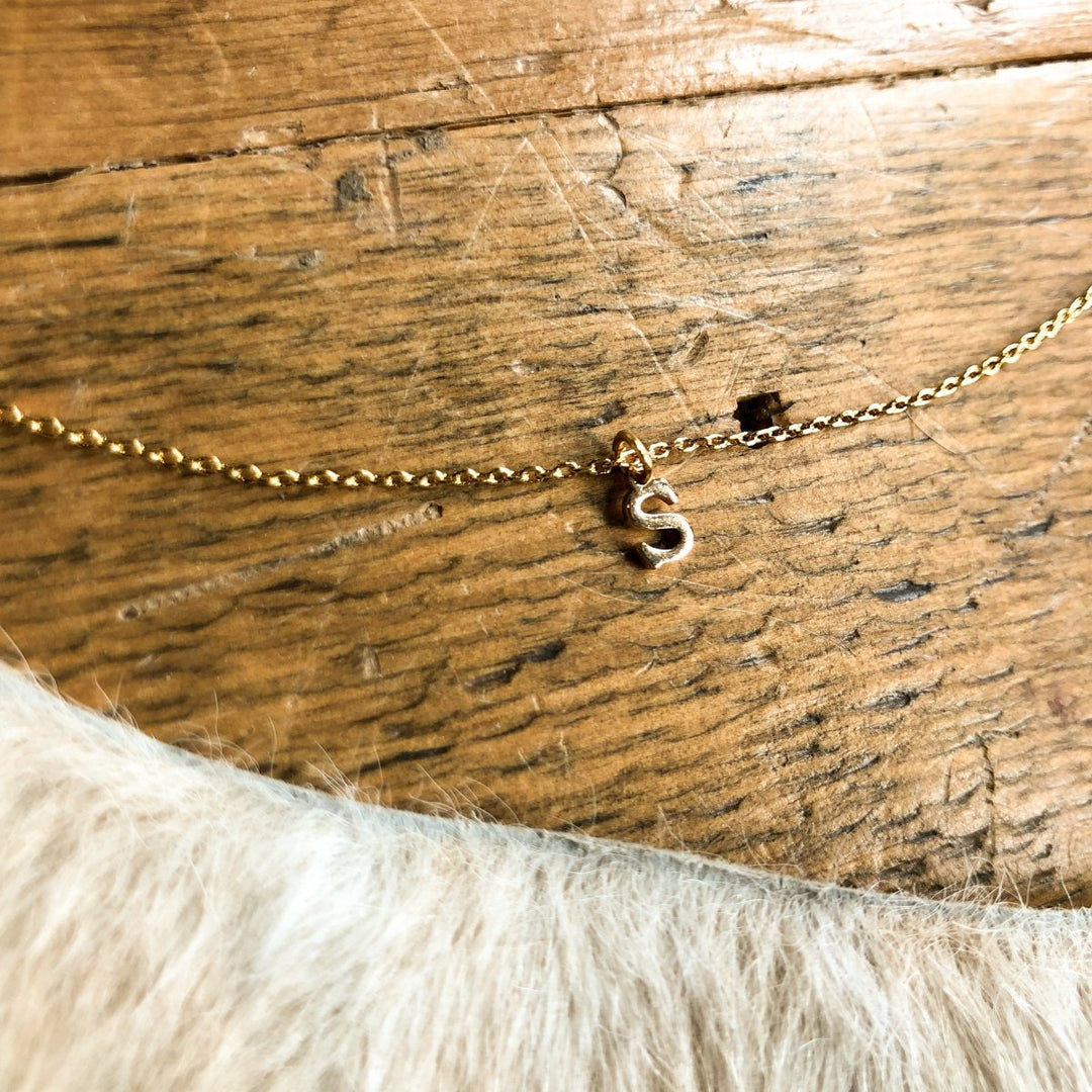 The Sylvan Initial Necklace by Savannah Sevens western life{&}style, featuring a gold S charm, rests on a wooden surface with a soft, furry object visible at the bottom of the image.