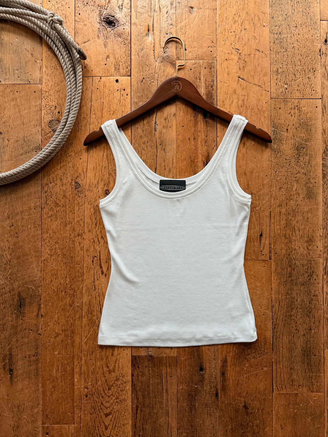Taft White Basic Tank with a ribbed scoop-neck hanging on a wooden hanger against a wooden floor with a coiled rope in the corner.