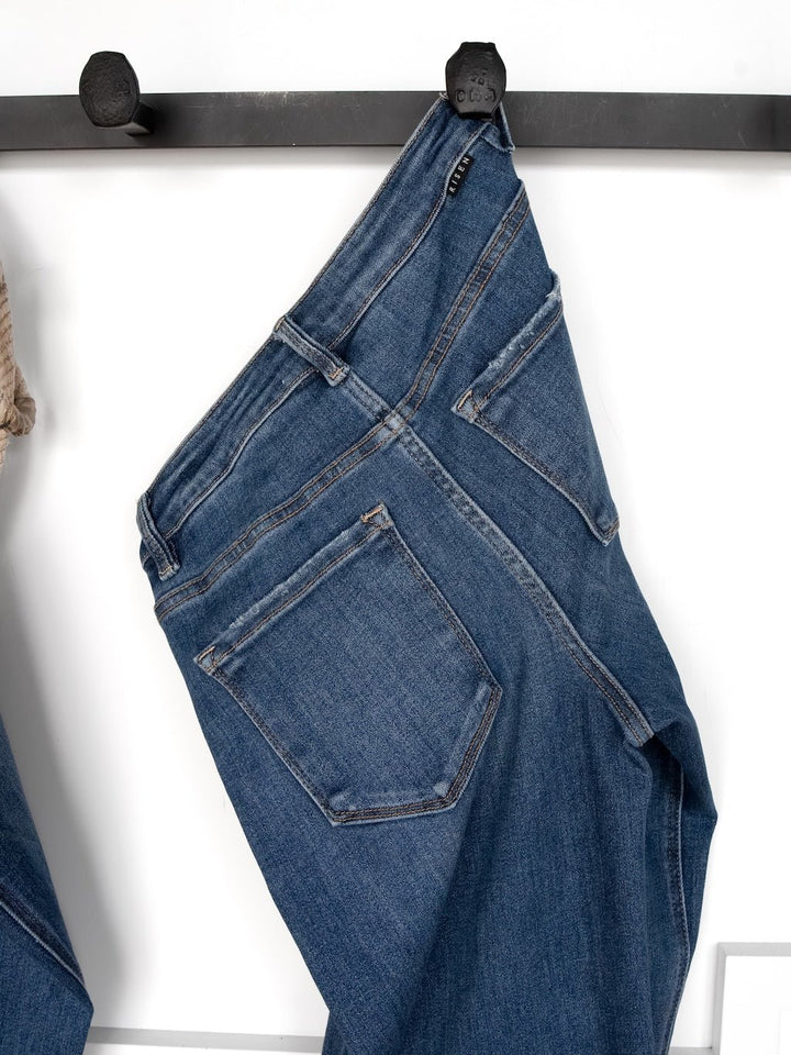 The Tagan High Rise Relaxed Straight Leg Jean by Savannah Sevens western life{&}style, in a medium dark wash, hangs by the belt loop on a black hook. This relaxed fit features visible back pockets and detailed stitching.