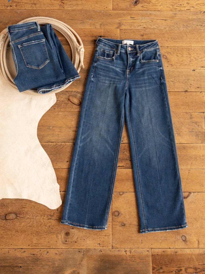 Two pairs of blue denim jeans from Savannah Sevens western life{&}style rest on a wooden floor: one folded in a rope, the other flat by a cowhide rug. Shown is the Tagan High Rise Relaxed Straight Leg Jean in medium dark wash.