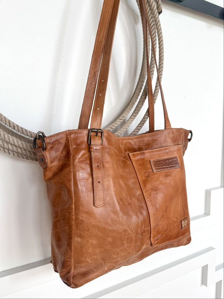 A Savannah Sevens western life{&}style Tahoka Leather Handbag in pecan dip dye with adjustable straps hangs on a decorative rope against a white wall. The bag features a front pocket and embossed logo patches.