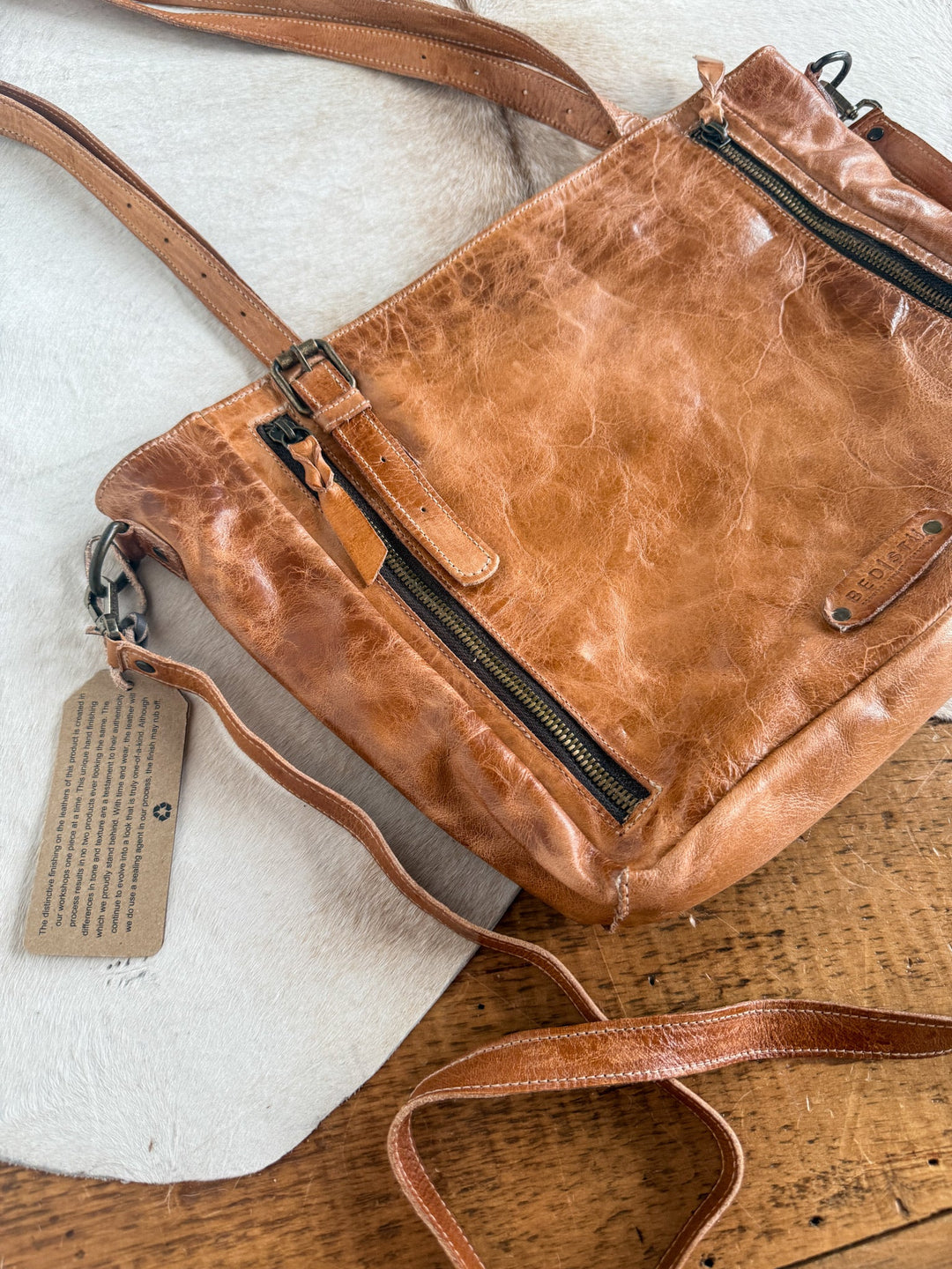 A Savannah Sevens western life{&}style Tahoka Leather Handbag, featuring a pecan dip dye finish, black zippers, and a shoulder strap, rests on a wooden surface and white fur. A tag with text is attached to one of the straps.