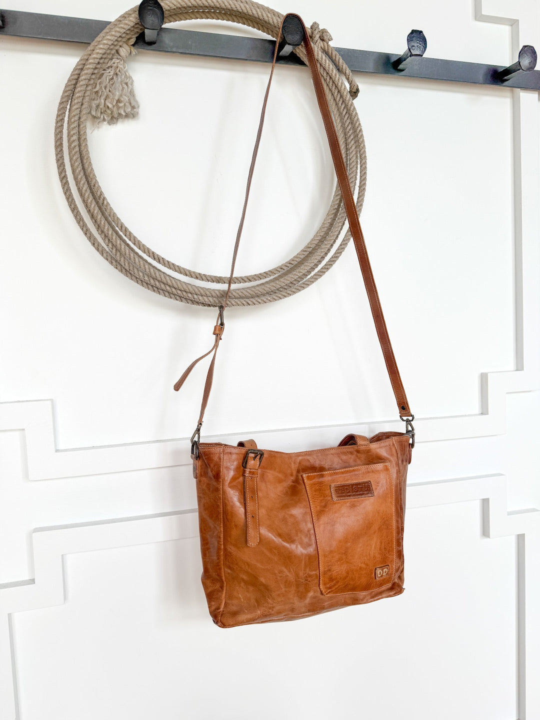 A Savannah Sevens western life{&}style Tahoka Leather Handbag hangs on a hook beneath a coiled rope on a white wall with decorative paneling.