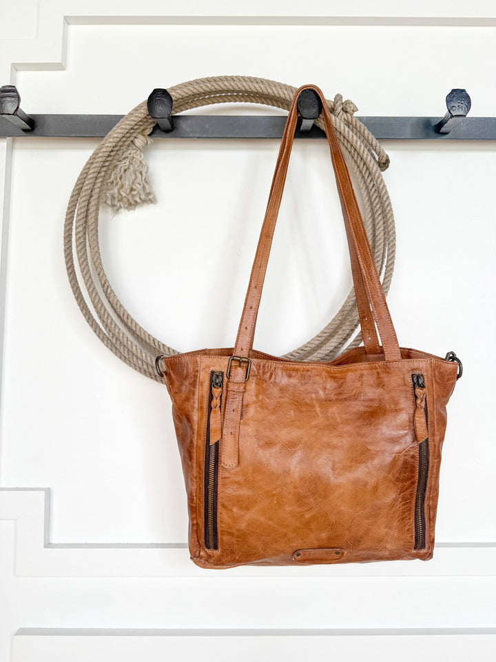 A Tahoka Leather Handbag by Savannah Sevens western life{&}style, featuring a pecan dip dye finish and two front zippers, hangs from a hook with a coiled rope beside it against a white paneled wall.
