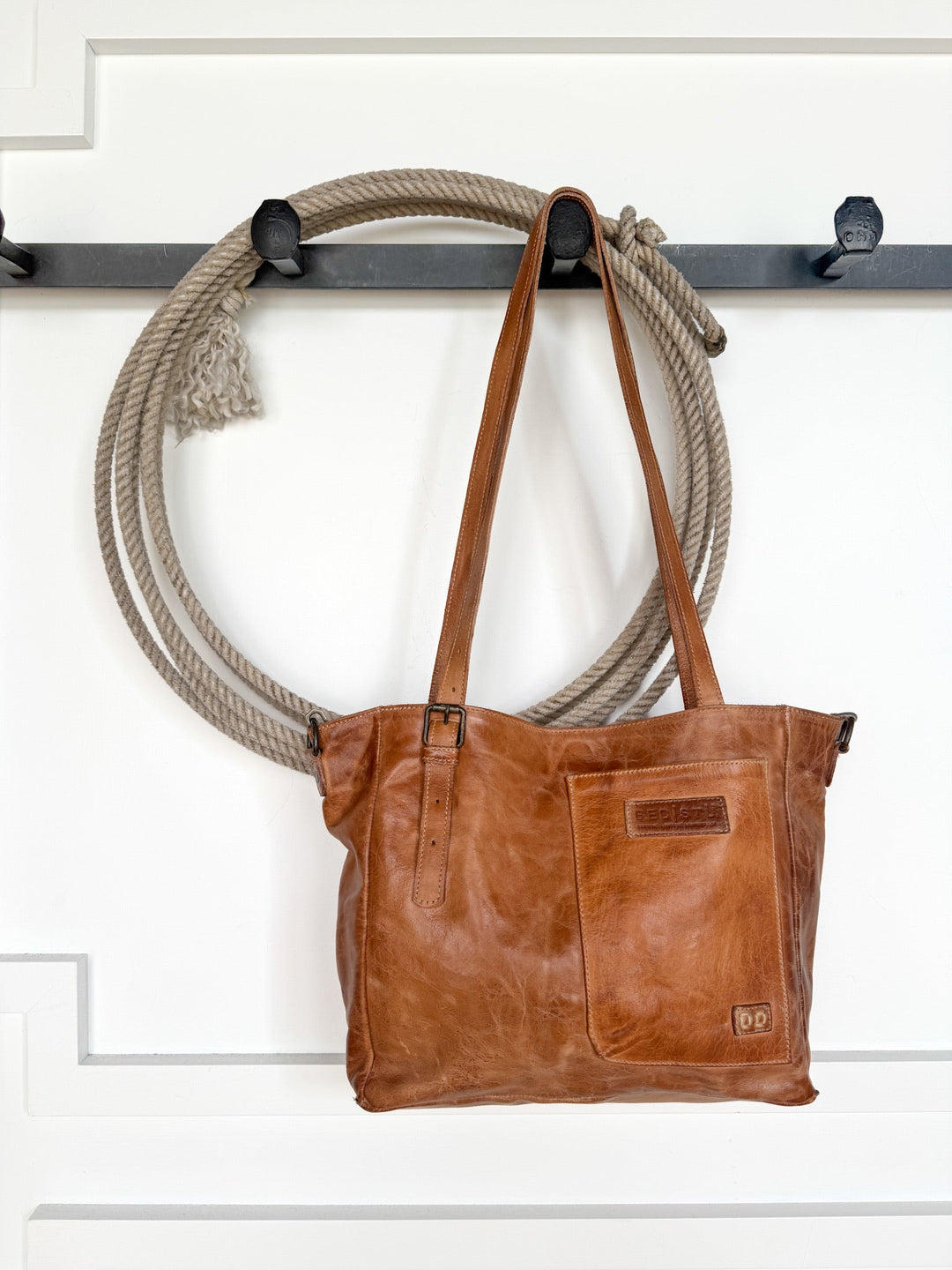 A Savannah Sevens western life{&}style Tahoka Leather Handbag in pecan dip-dyed leather hangs on a black wall hook, alongside a coiled rope draped over the hook, set against a white paneled wall.