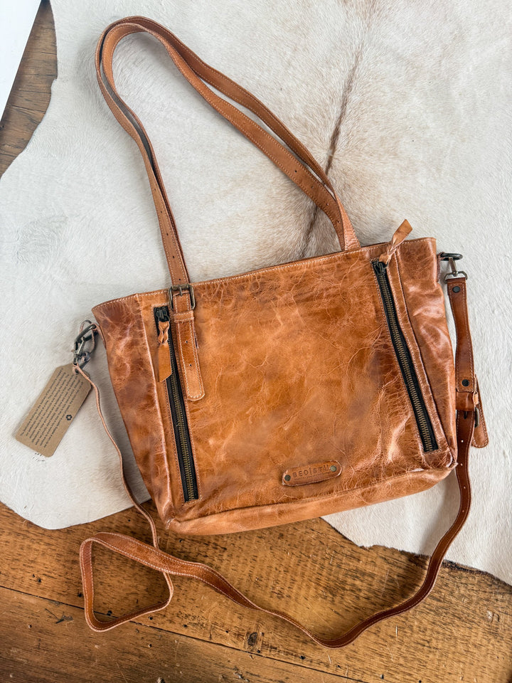 A Savannah Sevens western life{&}style Tahoka leather handbag in pecan dip dye, featuring two front zippers and long straps, rests on a white fur and wooden surface. A tan tag is attached to one strap.