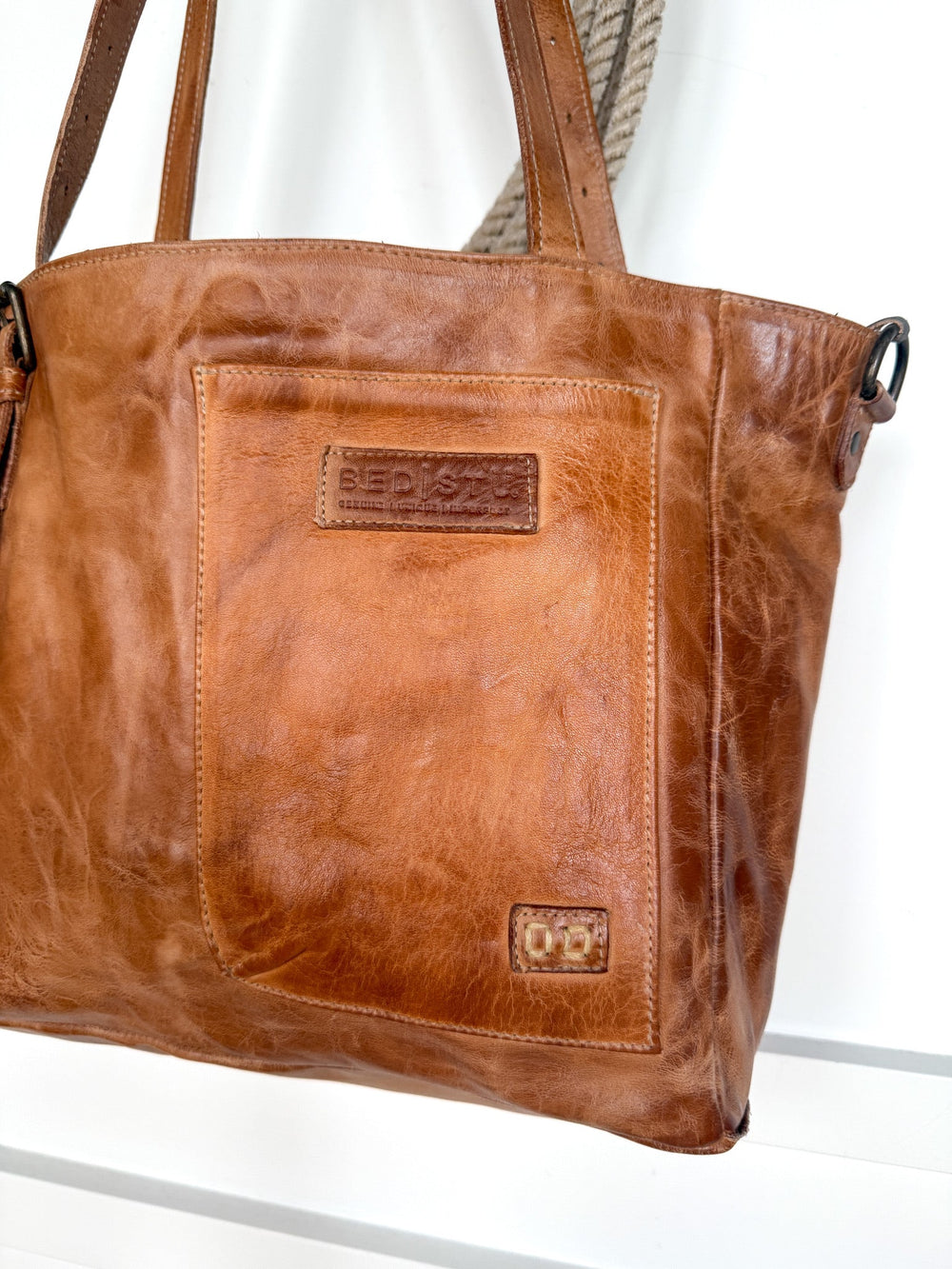 A pecan dip dye brown Tahoka Leather Handbag from Savannah Sevens western life{&}style, featuring a large front pocket and two short handles, hangs on a hook. The distressed leather finish is accented by two embossed brand patches on the front.