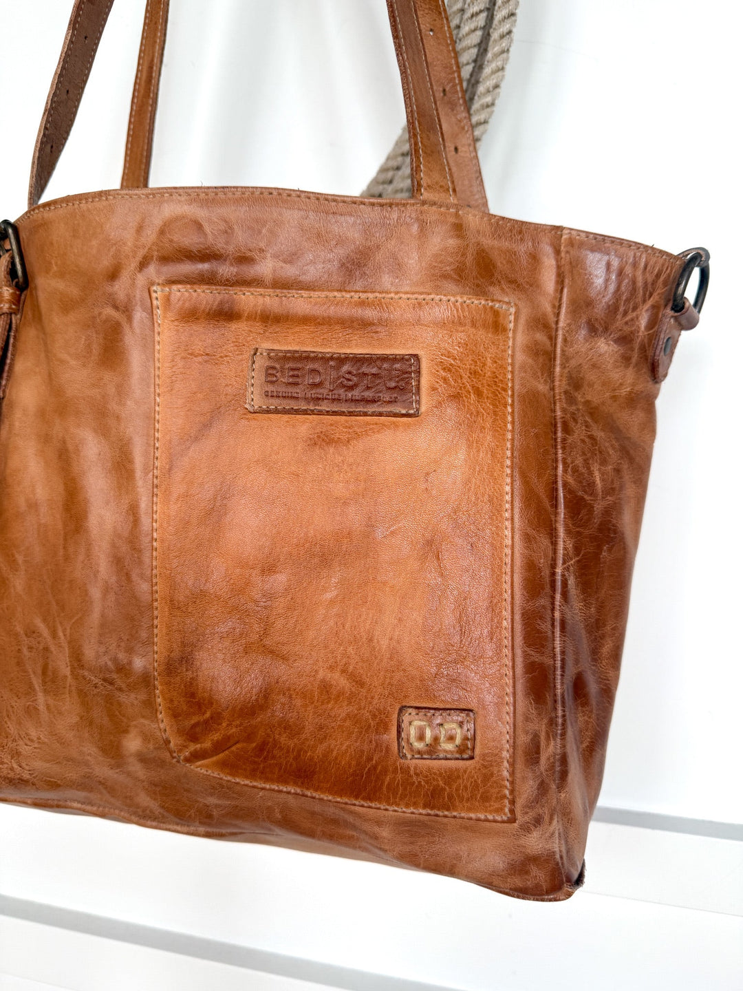 A pecan dip dye brown Tahoka Leather Handbag from Savannah Sevens western life{&}style, featuring a large front pocket and two short handles, hangs on a hook. The distressed leather finish is accented by two embossed brand patches on the front.