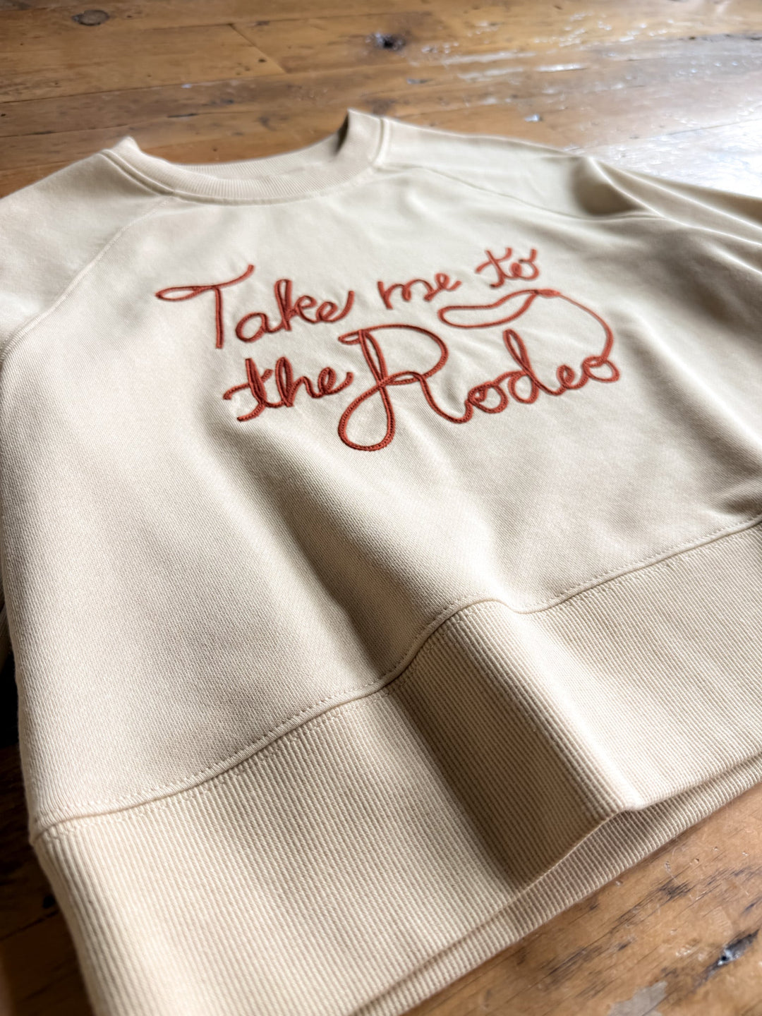 The “Take Me To The Rodeo Western Pullover” by Savannah Sevens western life{&}style, featuring red cursive chain stitch embroidery across the beige fabric and a relaxed fit, is displayed on a wooden surface.