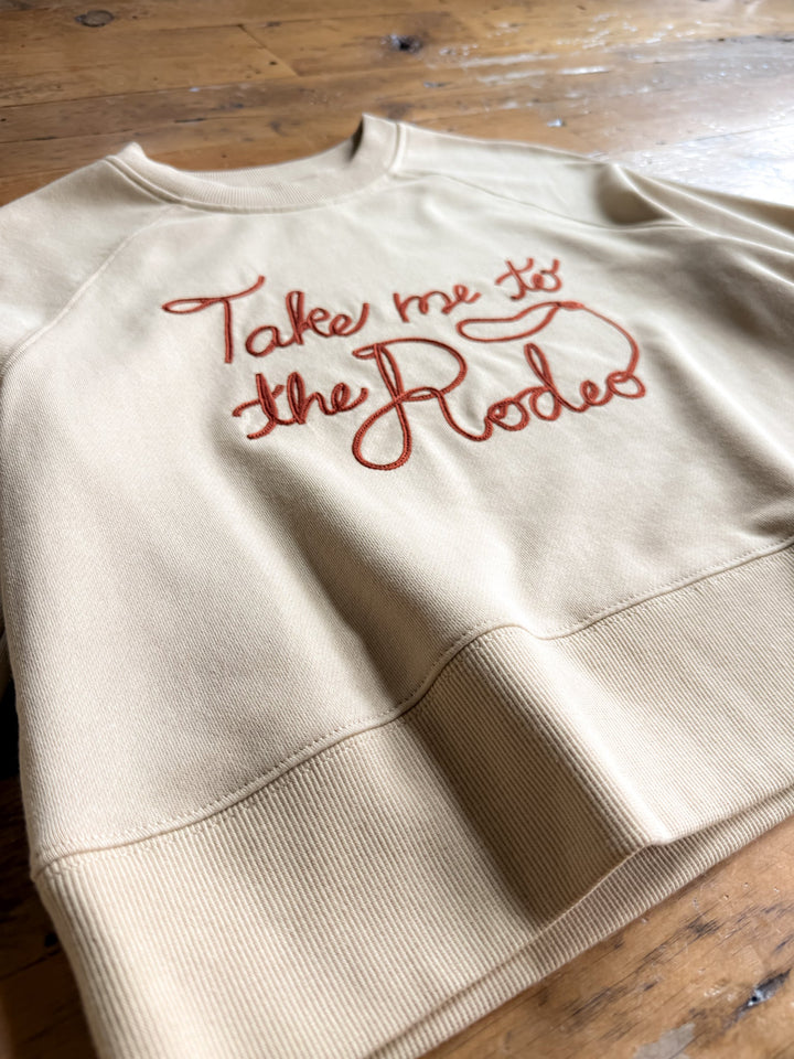 The “Take Me To The Rodeo Western Pullover” by Savannah Sevens western life{&}style, featuring red cursive chain stitch embroidery across the beige fabric and a relaxed fit, is displayed on a wooden surface.