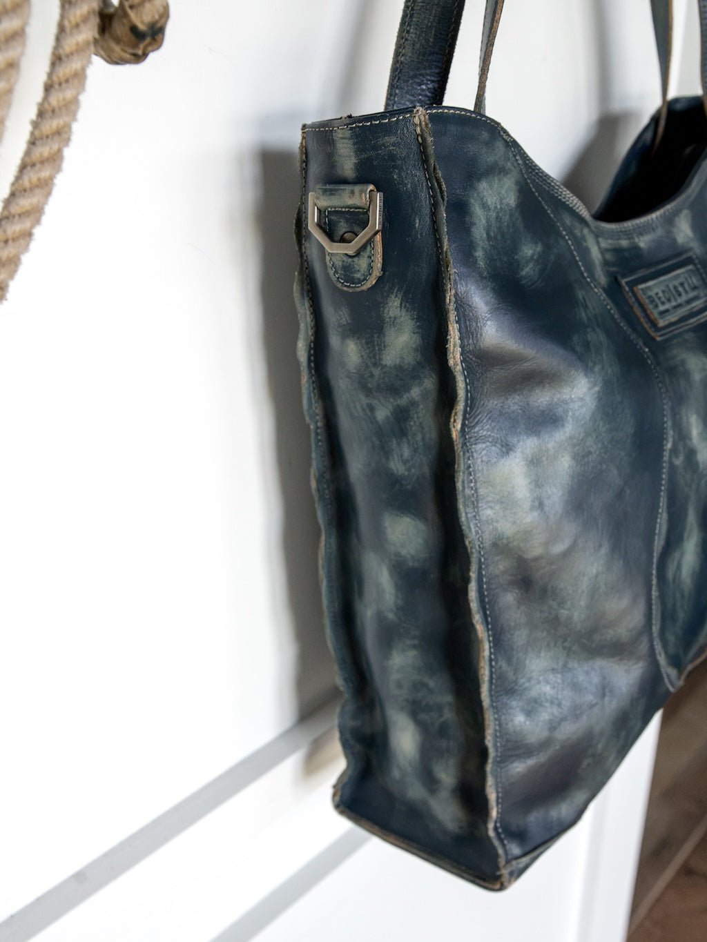 A close-up of the Tallmadge Leather Handbag by Savannah Sevens western life{&}style, showcasing its distressed dark blue leather, visible stitching, and worn textures as it hangs on a hook.