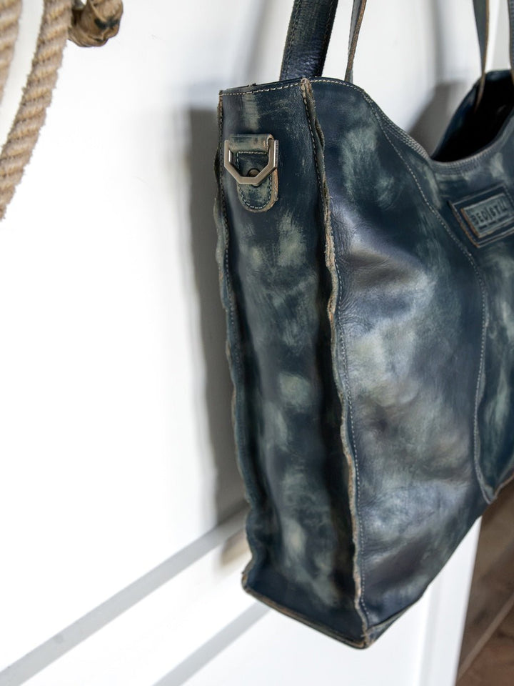 A close-up of the Tallmadge Leather Handbag by Savannah Sevens western life{&}style, showcasing its distressed dark blue leather, visible stitching, and worn textures as it hangs on a hook.