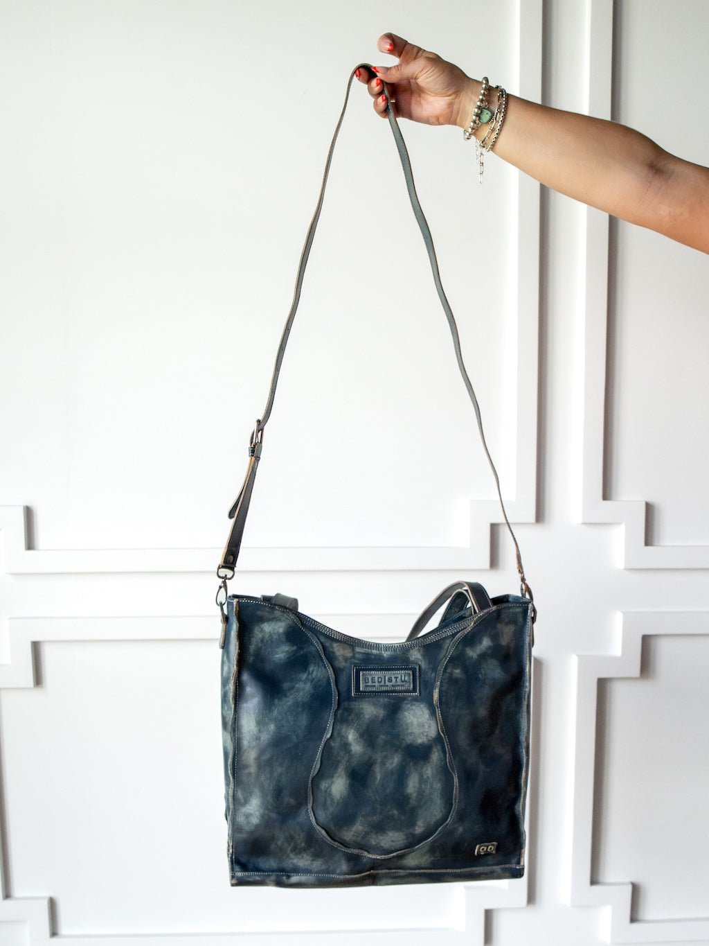 Wearing bracelets, a person holds up the Savannah Sevens western life{&}style Tallmadge Leather Handbag in blue-gray by its long crossbody strap against a white, decorative panel wall.