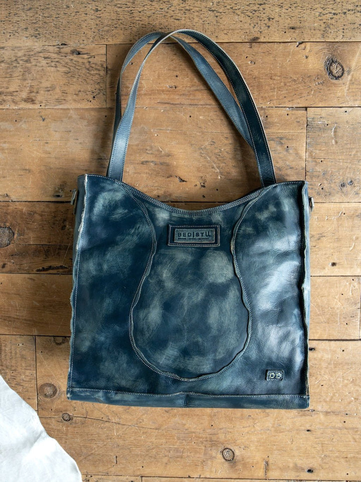 The Tallmadge Leather Handbag from Savannah Sevens western life{&}style, in blue leather, sits on a wooden surface. This tote features double handles, a crossbody strap, visible stitching, a front pocket, and a rectangular logo patch near the top.