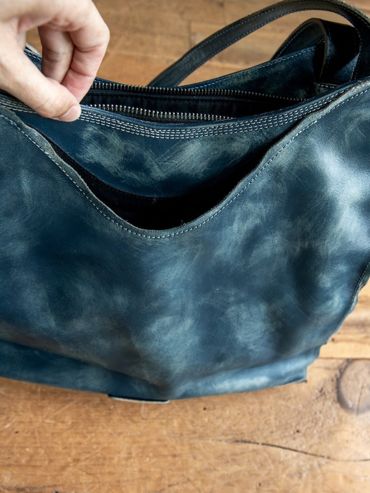 A hand holds open the zipper of the Tallmadge Leather Handbag by Savannah Sevens western life{&}style, a dark, worn leather bag featuring a front pocket and a crossbody strap, resting on a wooden surface.