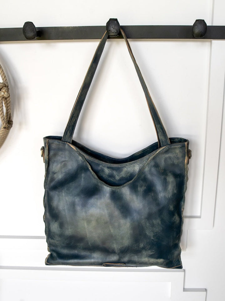 A large Tallmadge Leather Handbag by Savannah Sevens western life{&}style, in dark blue leather with two straps, hangs on a black wall hook against a white paneled background.