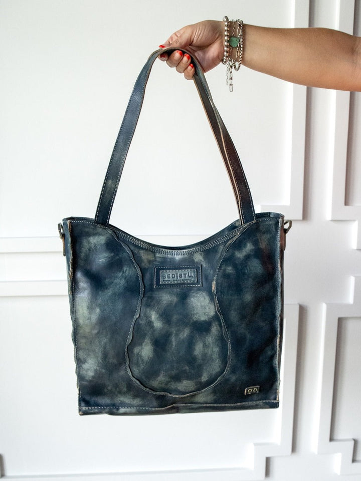 A hand adorned with bracelets holds the Tallmadge Leather Handbag by Savannah Sevens western life{&}style, showcasing its distressed dark leather, visible stitching, branded Bedstu label, and crossbody strap details against a white textured wall.