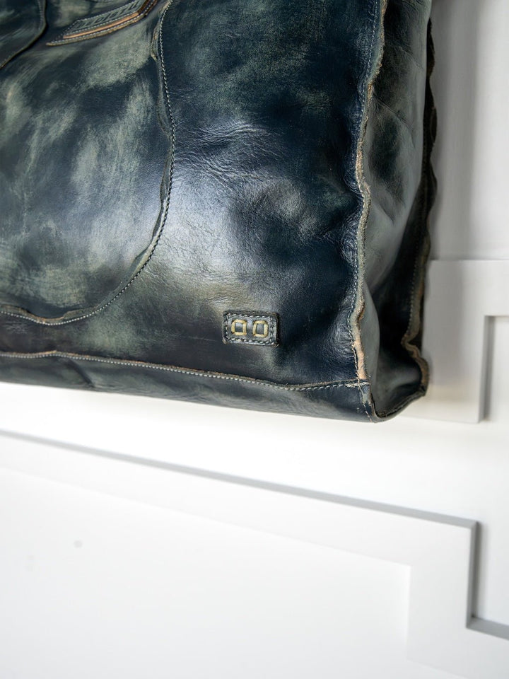 A close-up of the Tallmadge Leather Handbag by Savannah Sevens western life{&}style showcases its dark, distressed leather with visible stitching and a small metallic square accent. The crossbody strap is also featured, all set against a white background with geometric molding.