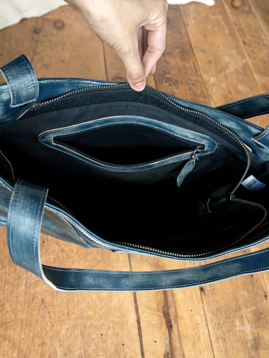 A hand holds open the Tallmadge Leather Handbag by Savannah Sevens western life{&}style, showcasing its blue leather exterior with a zippered inner pocket and a spacious black fabric-lined interior, all set against a wooden floor. This elegant handbag seamlessly blends style and functionality.